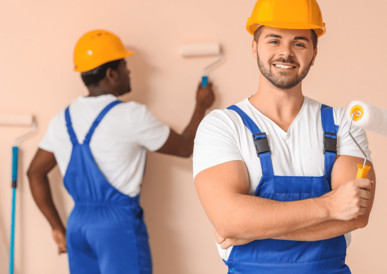 Smiling commercial painters in blue overalls and yellow helmets, painting a wall with rollers in a professional setting.